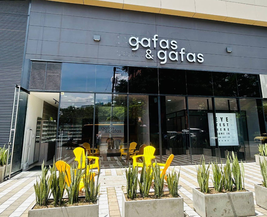 Glass-front storefront for 'gafas & gafas' with bright yellow outdoor chairs and potted plants outside. A sunny urban sidewalk scene inside shows more displays.